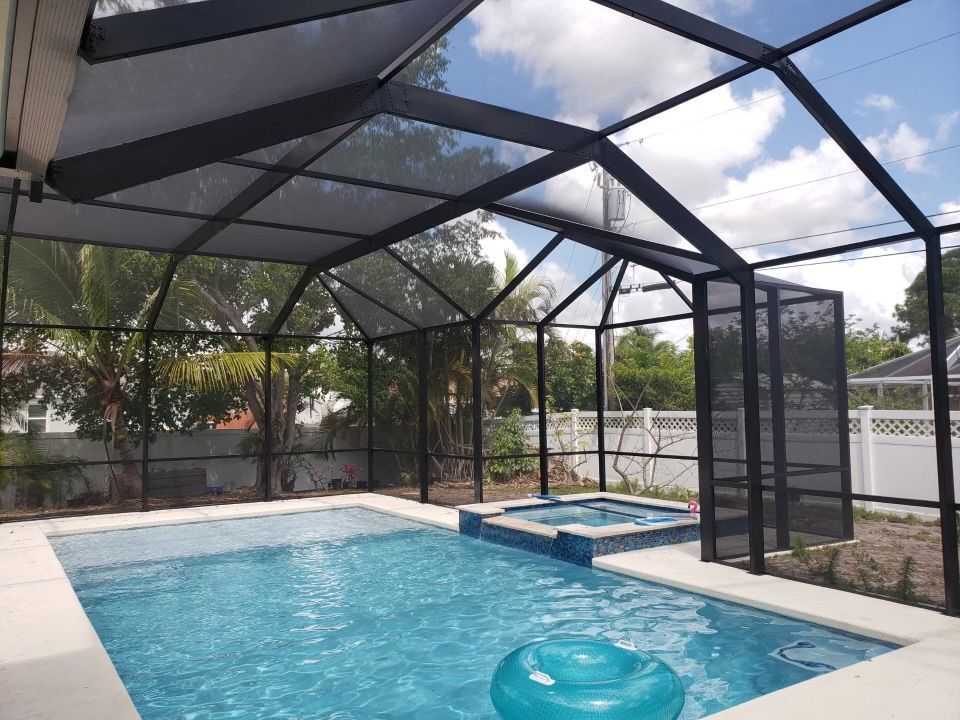 Pool enclosed by black screening under a sunny sky. Blue water, white tile, and a hot tub.