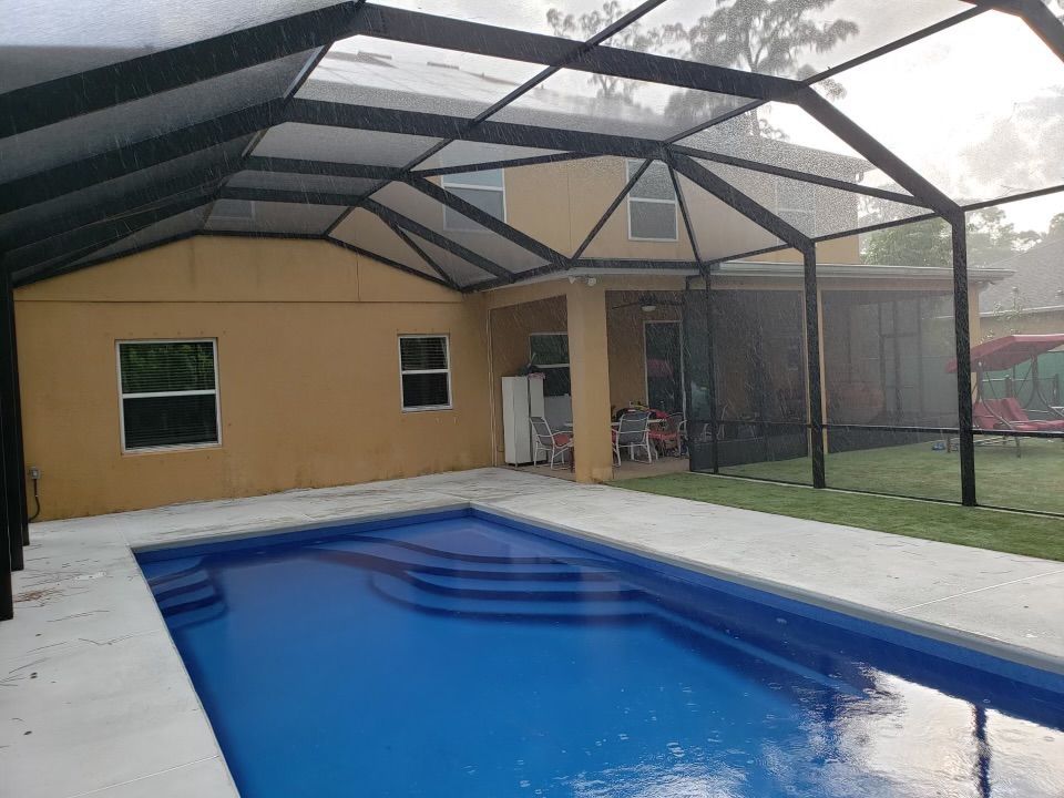 Swimming pool under a screened enclosure attached to a light brown house; overcast day.