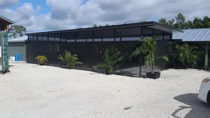 Black building with screen walls, palm trees, and gravel parking area. Bright sunny day.