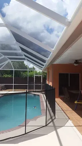 A screened-in pool area with blue water, surrounded by a black safety fence, under a cloudy sky.