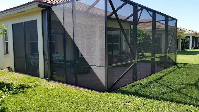 Screened-in patio enclosure attached to a beige house, surrounded by green grass and a sunny sky.