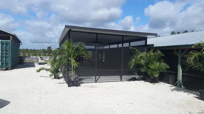 A screened enclosure with a dark roof and walls, flanked by potted palm trees, on a gravel lot under a cloudy sky.