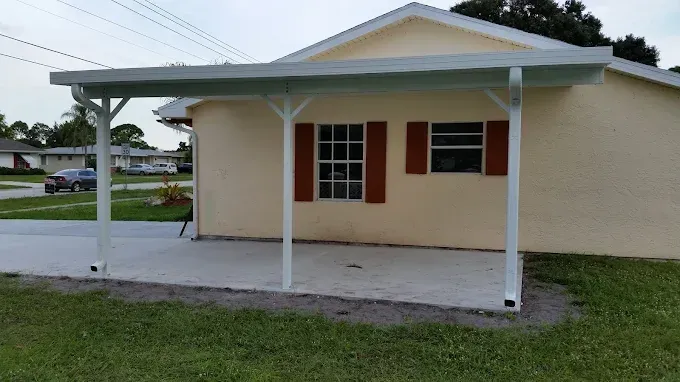 Carport attached to a light yellow house with red shutters. Green grass in front, car in the distance.