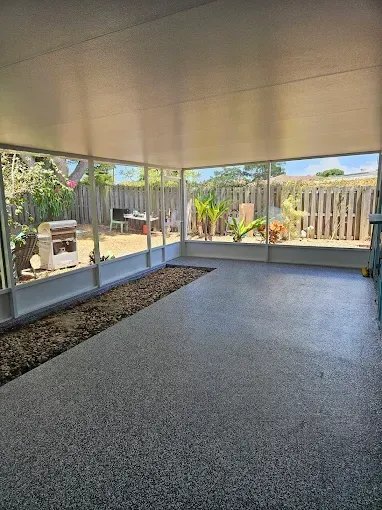 Enclosed patio with gray flooring, gravel bed, and views of a backyard with a grill and trees.