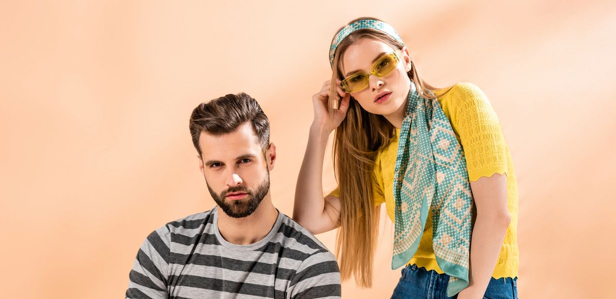 Un uomo con una camicia a righe grigie siede accanto a una donna con una maglietta gialla, una sciarpa blu a fantasia e occhiali da sole colorati.