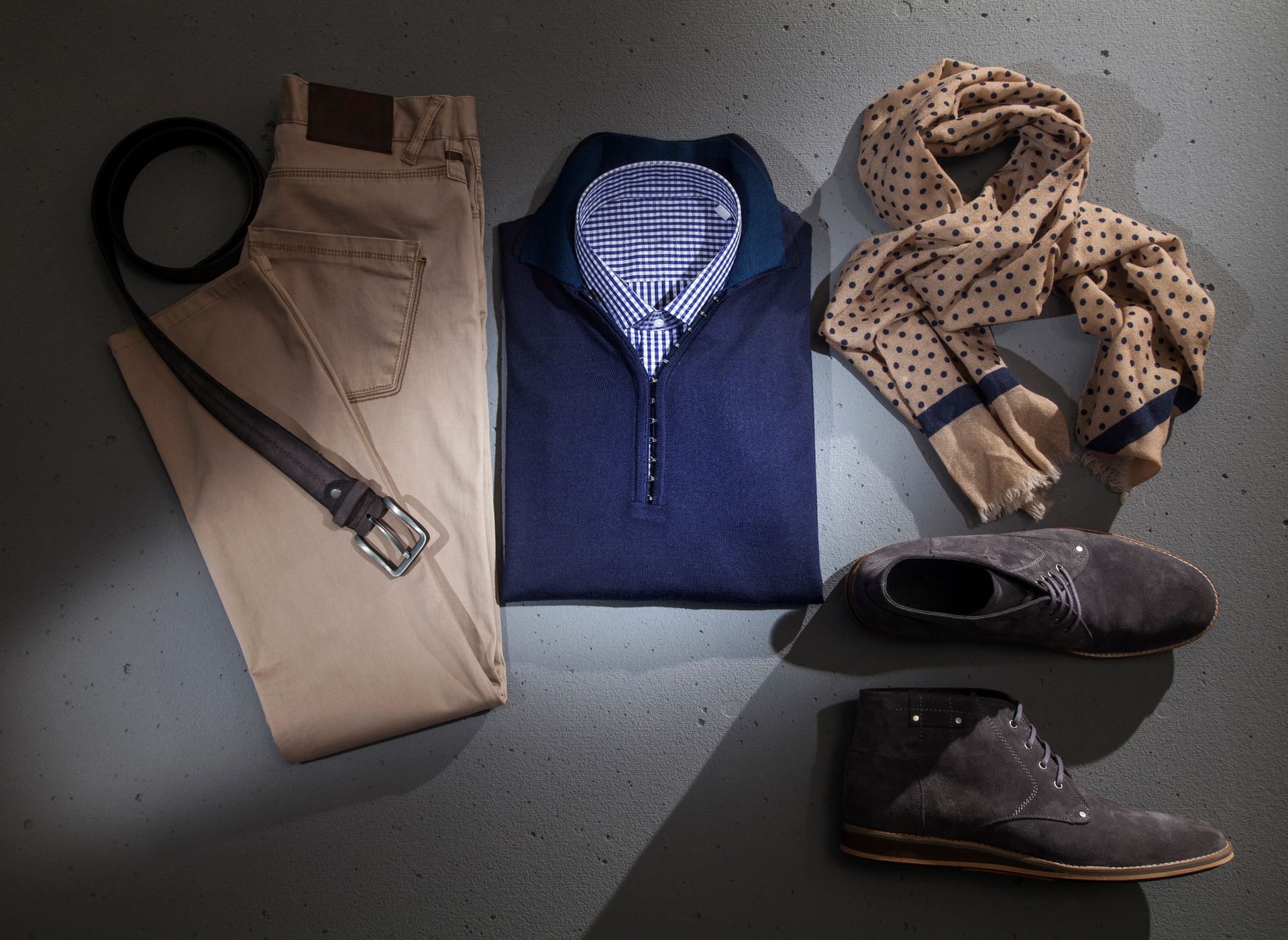 Una composizione di abbigliamento maschile: pantaloni color kaki, maglione blu scuro sopra una camicia a quadri, sciarpa a pois e scarpe scamosciate grigie.