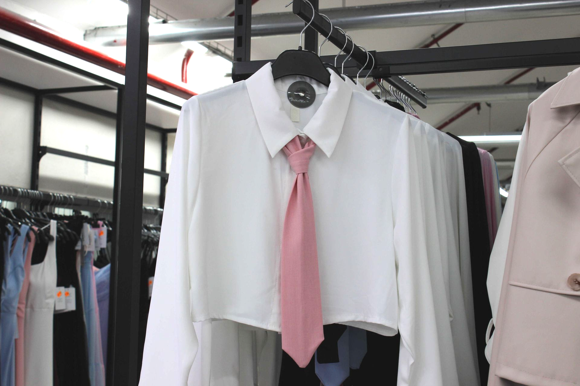 Una camicia bianca corta con bottoni, abbinata a una cravatta rosa, esposta su una gruccia in un negozio di abbigliamento.