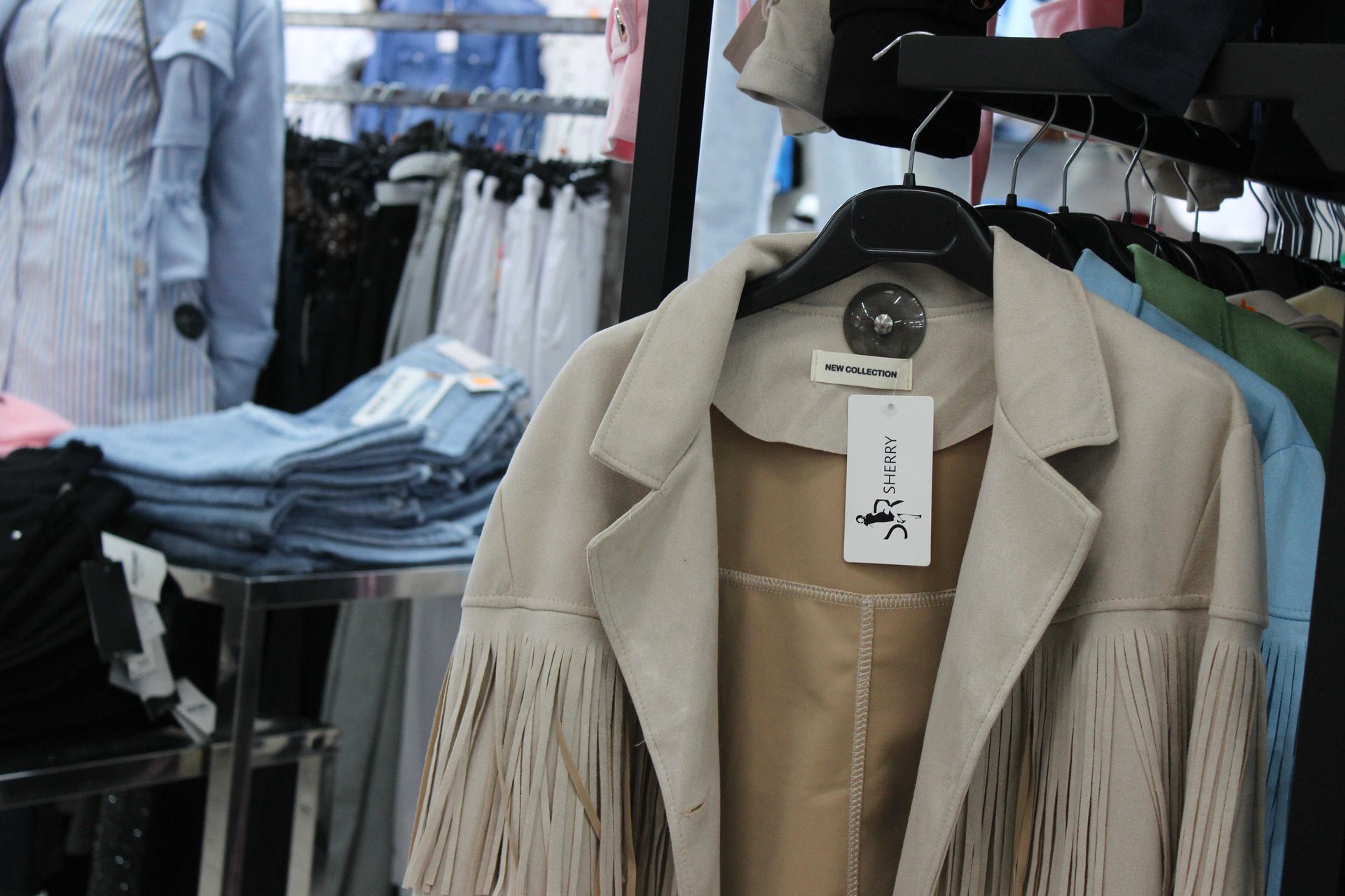 In un negozio di abbigliamento, una giacca beige chiaro con frange è appesa a uno stand, con jeans piegati e altri capi d'abbigliamento nelle vicinanze.