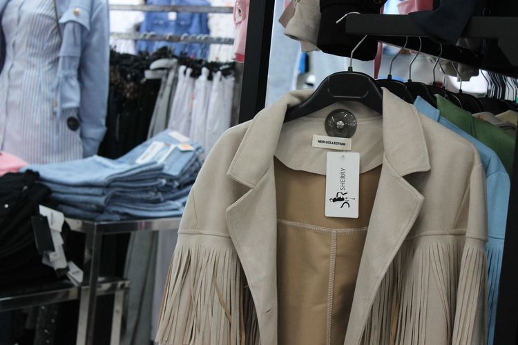 In un negozio di abbigliamento, una giacca beige chiaro con frange è appesa a uno stand, con jeans piegati e altri capi d'abbigliamento nelle vicinanze.