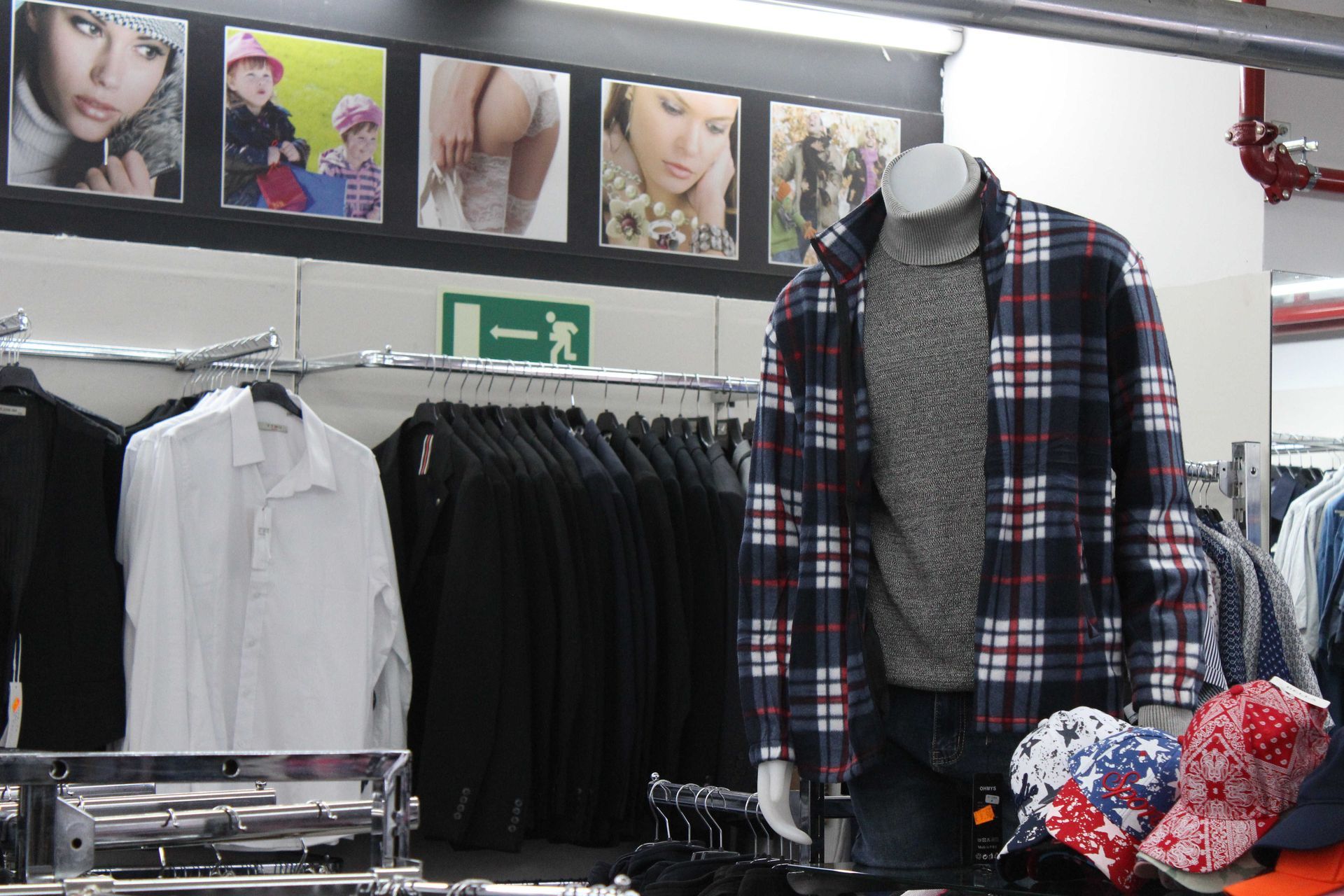 Vetrina di un negozio di abbigliamento con un manichino che indossa una giacca a quadri sopra un maglione, con camicie e cappelli sugli scaffali vicini.