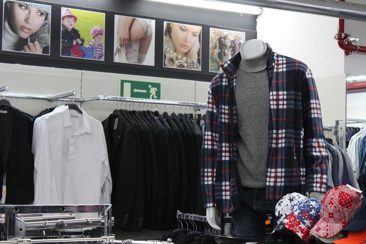 Vetrina di un negozio di abbigliamento con un manichino che indossa una giacca a quadri sopra un maglione, con camicie e cappelli sugli scaffali vicini.
