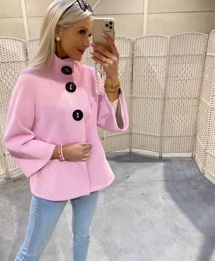 Una persona in piedi davanti a un paravento bianco, indossa una giacca a trapezio rosa chiaro a tre bottoni e jeans blu.