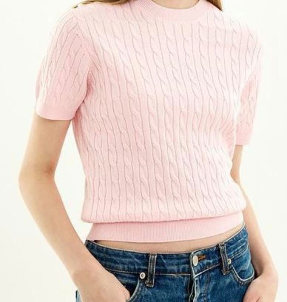 Persona che indossa una maglia a maniche corte rosa chiaro e jeans blu, in piedi davanti a uno sfondo bianco.