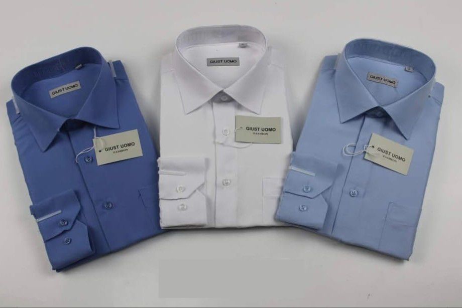 Tre camicie eleganti piegate – blu, bianca e azzurra – esposte su uno sfondo bianco.