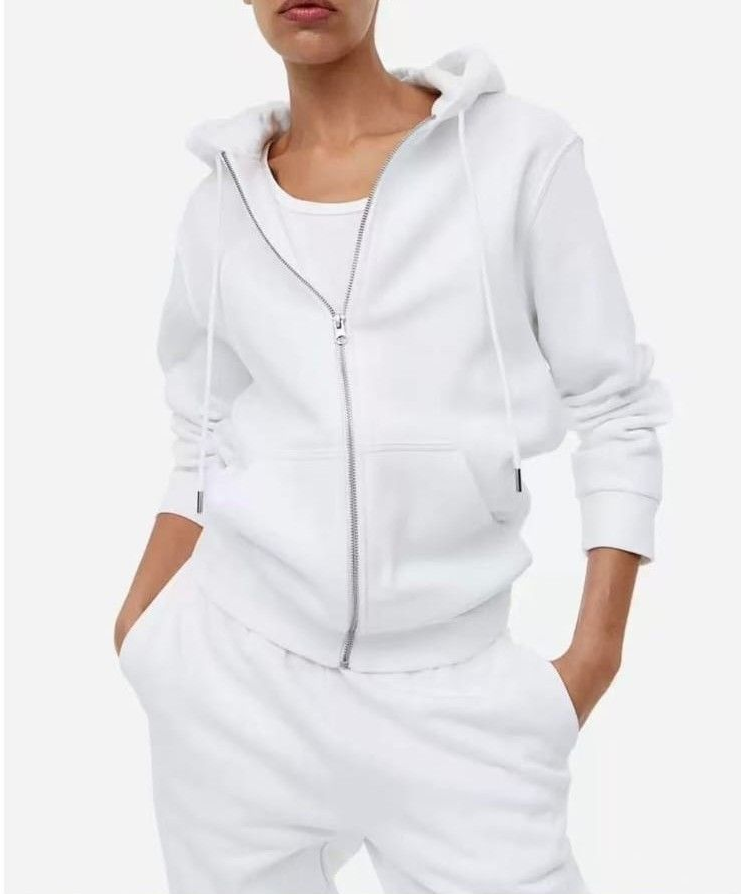 Donna con felpa bianca con zip e pantaloni della tuta, con le mani in tasca.