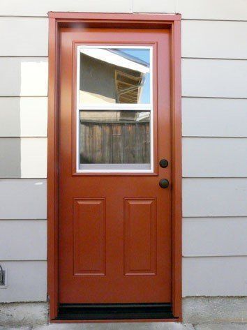 Brown exterior door with window, two panels, and black hardware; set in a red frame against gray siding.