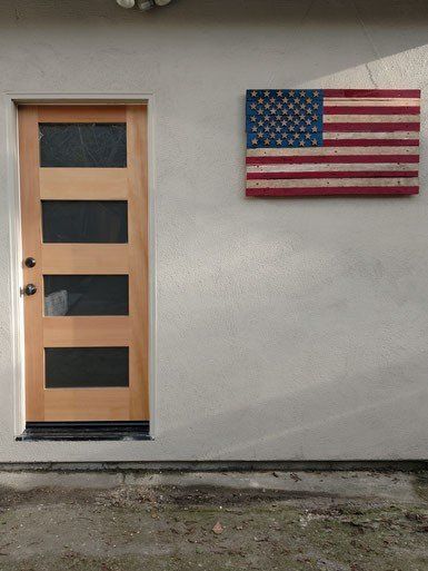 Wooden door with glass panels beside a weathered American flag on a textured, light gray wall.