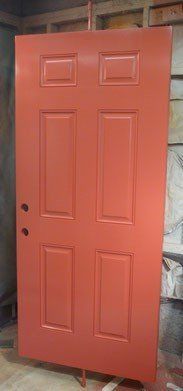 Orange painted six-panel door with door hardware, propped up in a workshop.