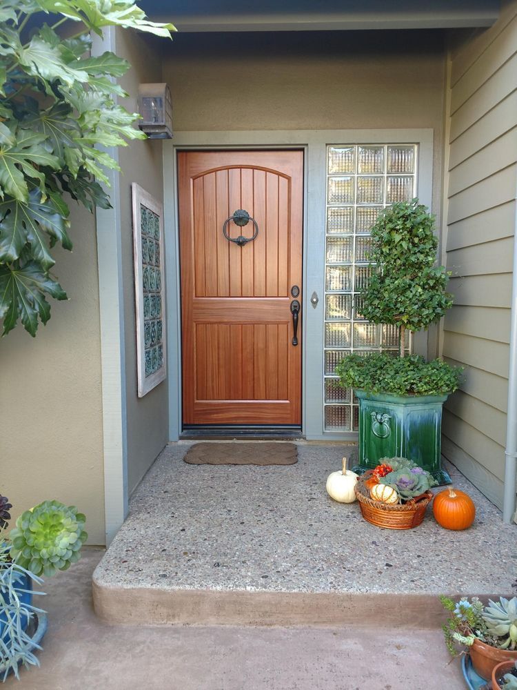 Wooden front door with glass block window, pumpkins, and a potted topiary on a concrete porch.