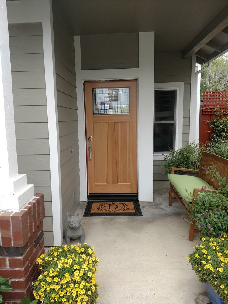 Wooden front door with glass panel, flanked by white pillars, on a porch with bench and flowers.