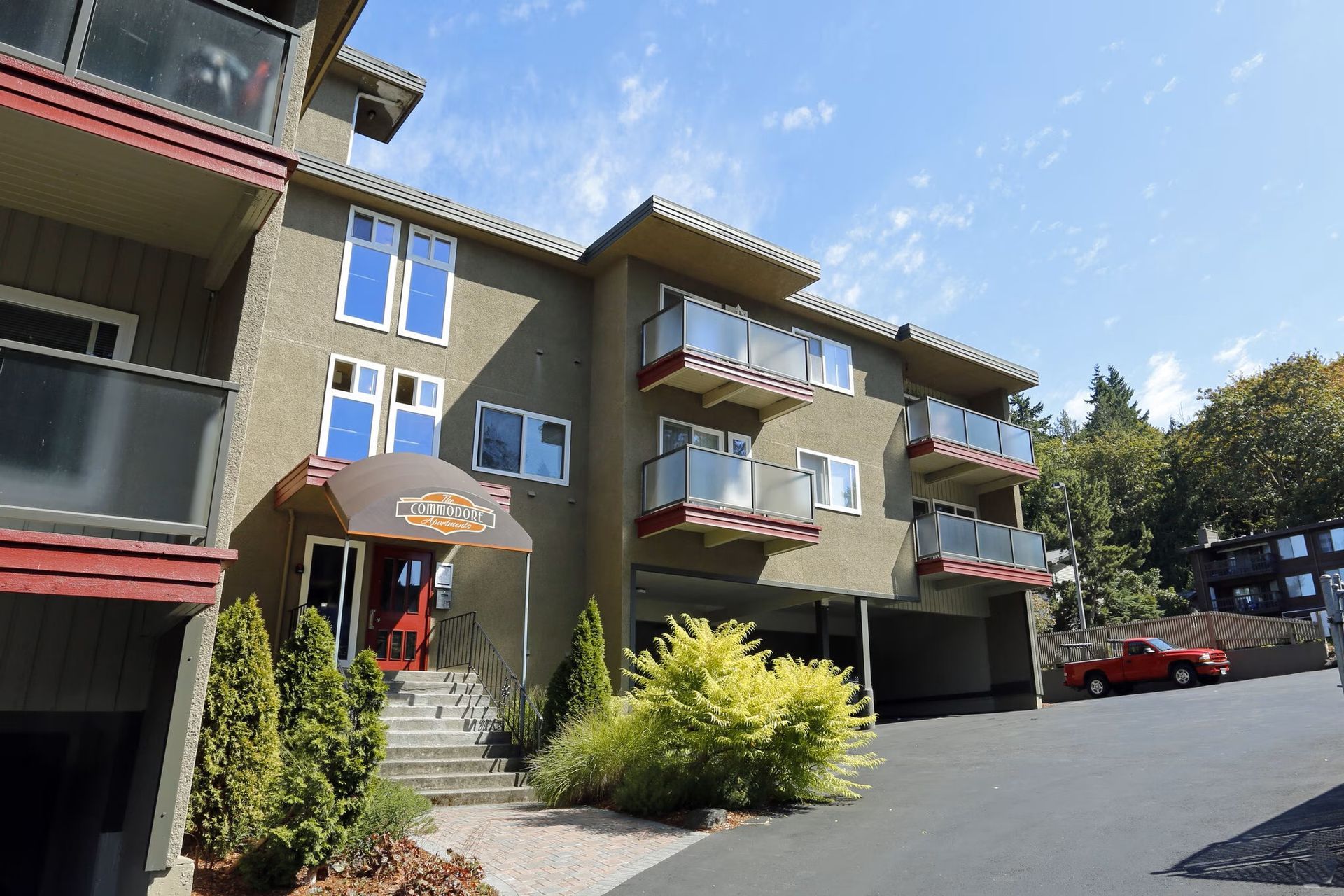 Apartment building with balconies and a red entrance door, set on a paved driveway.