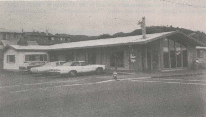 A vintage, low-slung building with large windows and cars parked in front, likely a business on a corner.