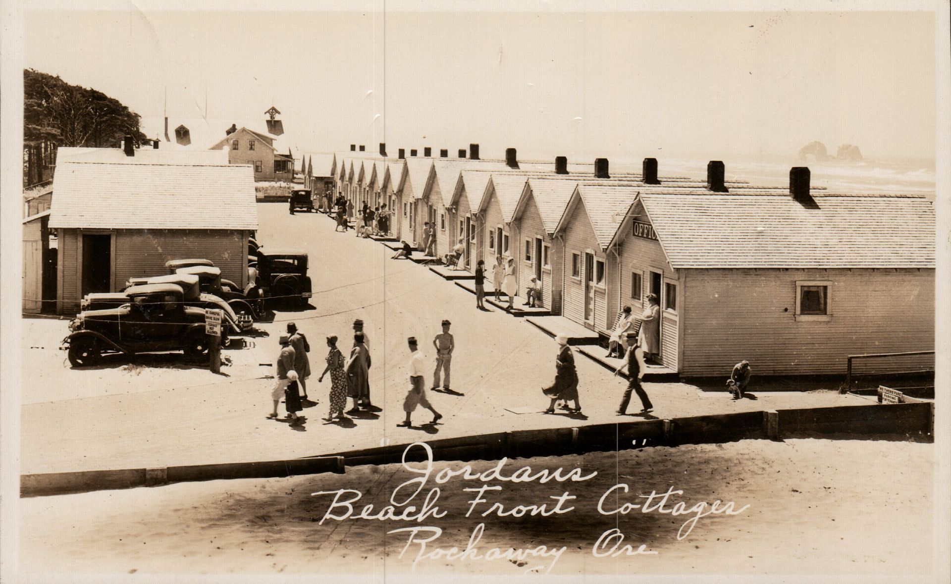 Row of beach cottages, people walking, cars parked. 
