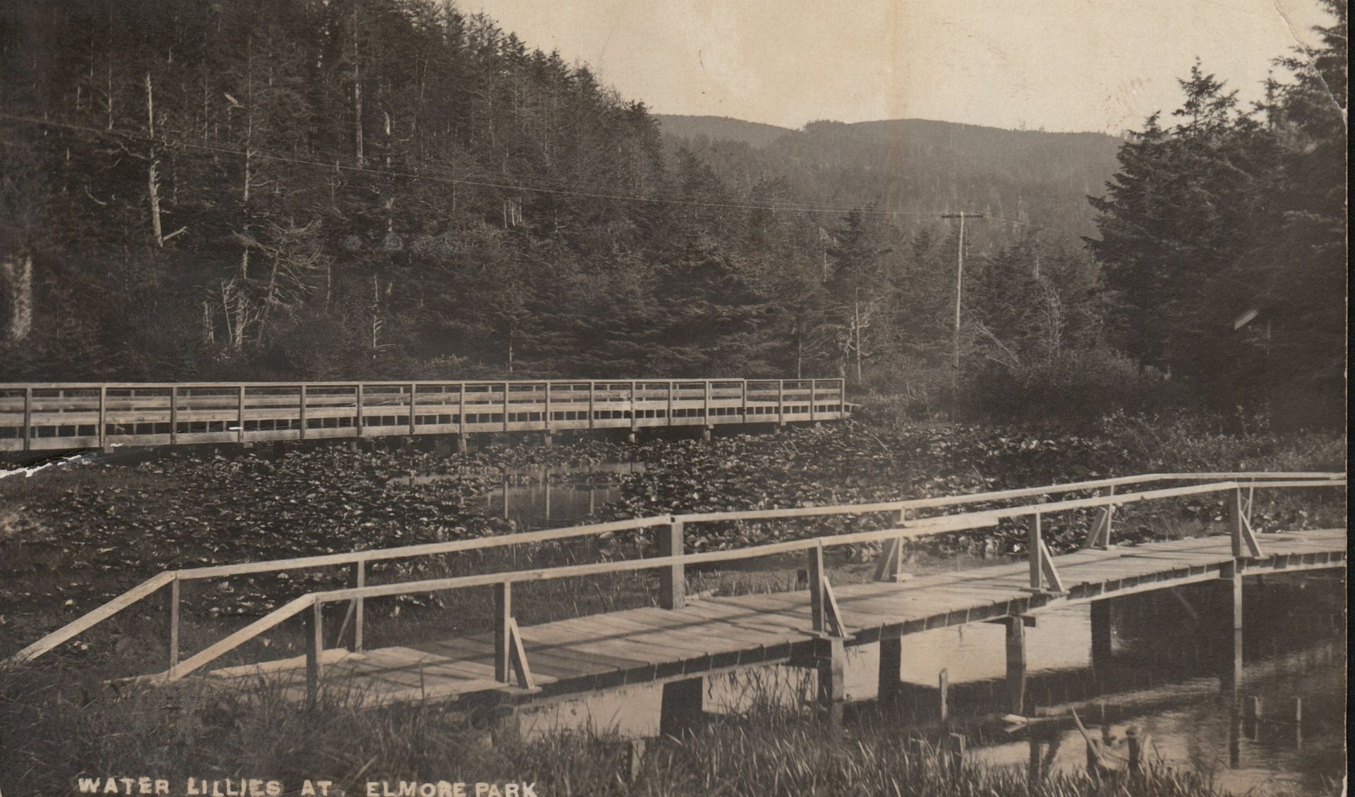 Sepia river scene with wooden bridges, forested hills, and a distant road railing.