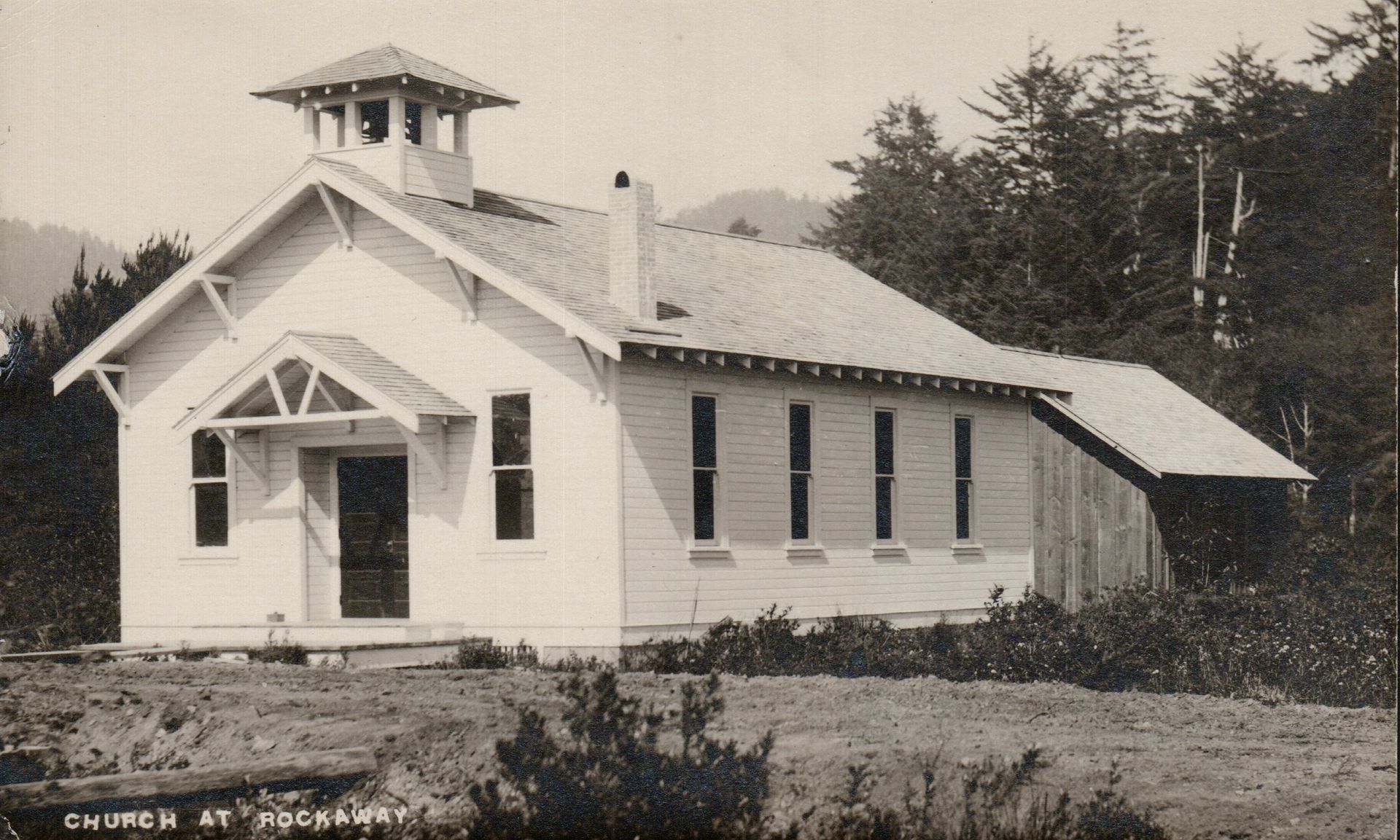 White clapboard church with bell tower in a grassy field, trees in the background.