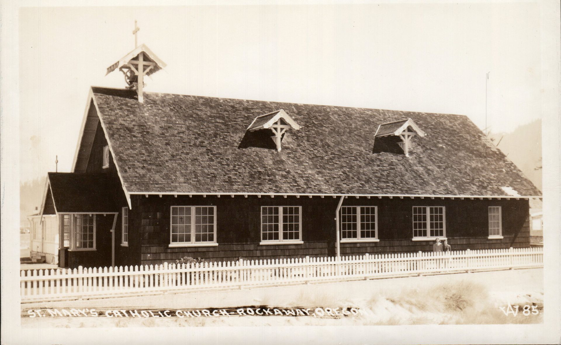 Black and white photo of a wood-shingled building with a white picket fence and small bell tower.