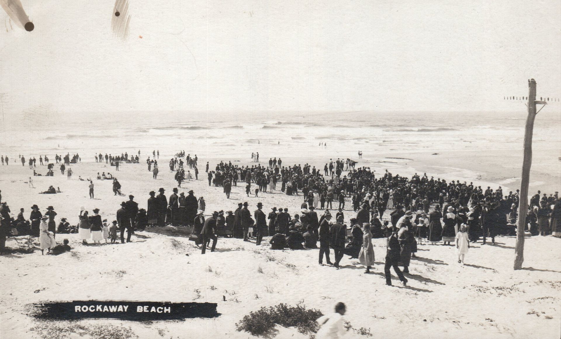 Large crowd gathered on a sandy beach. Ocean visible in the background.