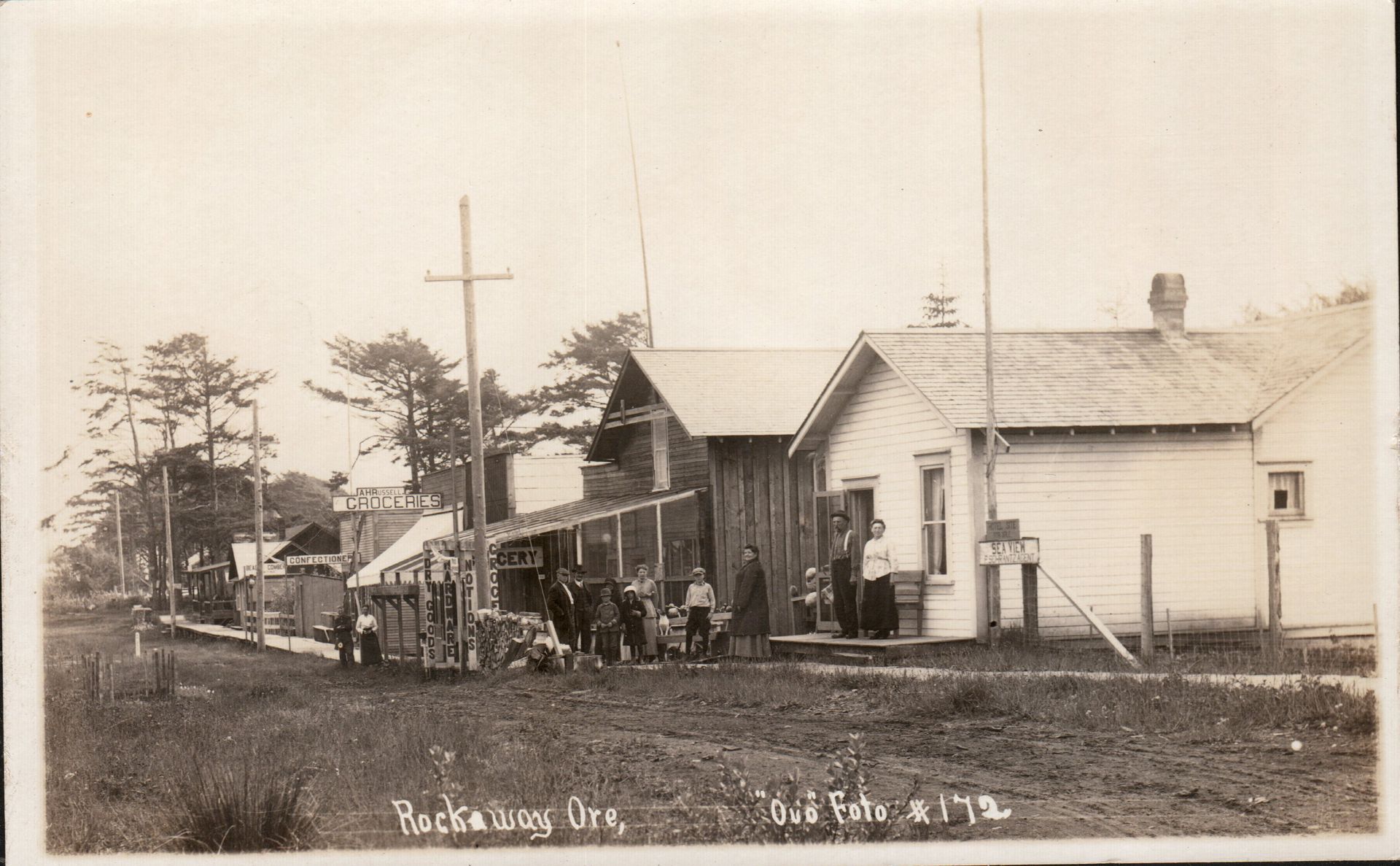 Sepia photo of a small village street with storefronts, people, trees, and a white house with a front porch.