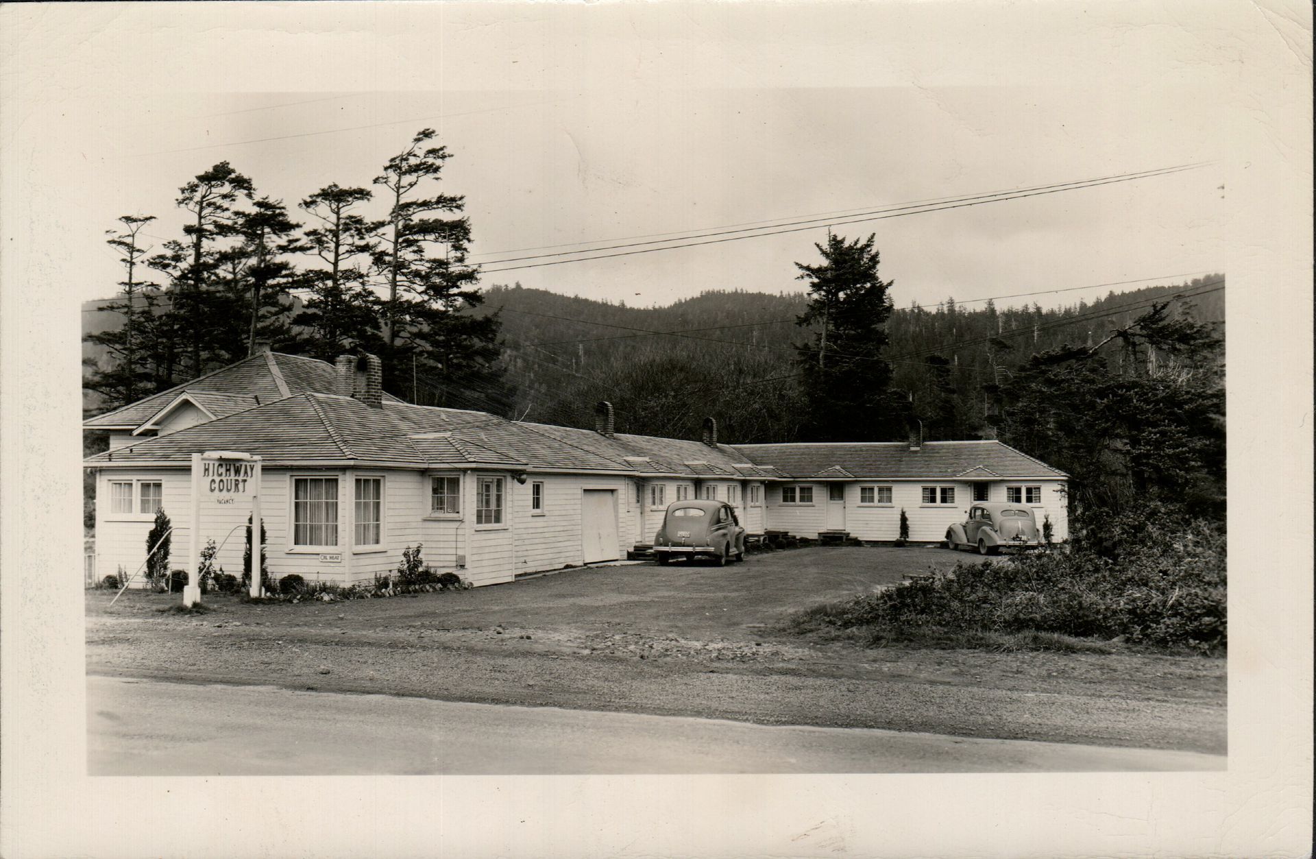 Motel with cars parked outside, trees and a mountain in the background.