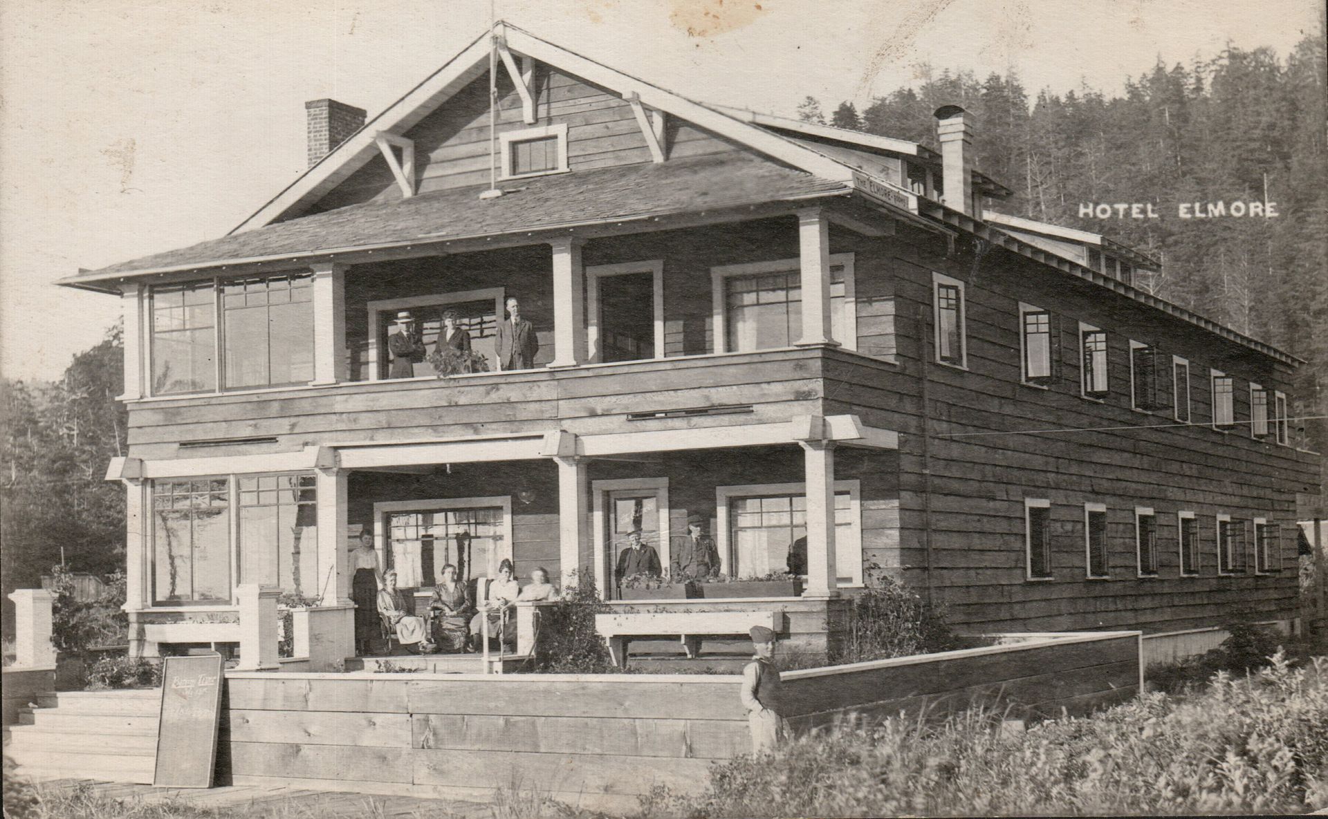 Hotel Elmore, two-story wooden building with porch and balcony; guests relax outside; forest background.