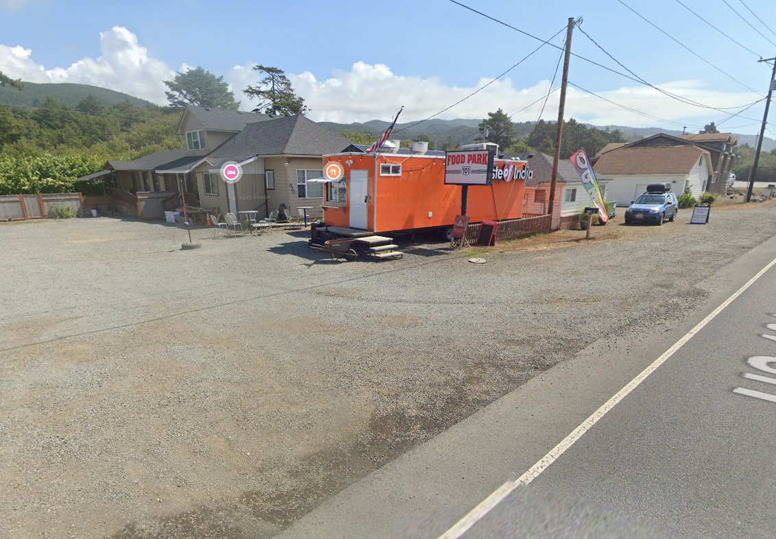 Orange food truck and house in gravel lot next to a road, surrounded by green hills and blue sky.