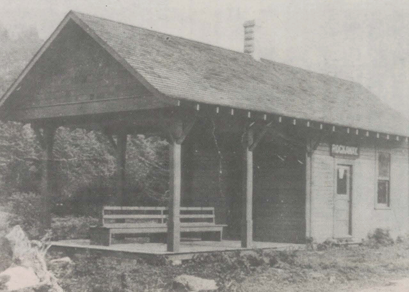 Black and white photo of a small train depot with a covered porch, benches, and a single door.
