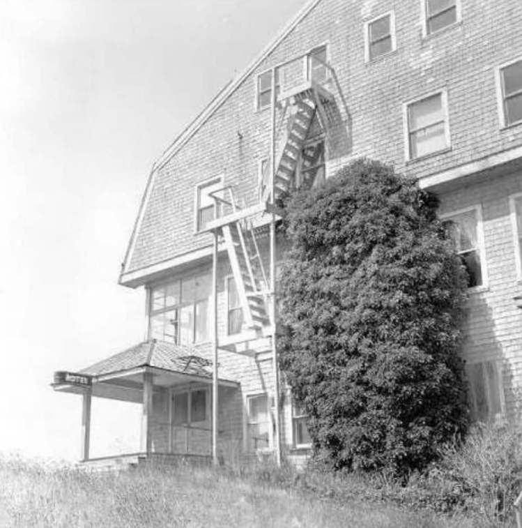 Black and white photo of a weathered motel with a fire escape and overgrown shrubbery.