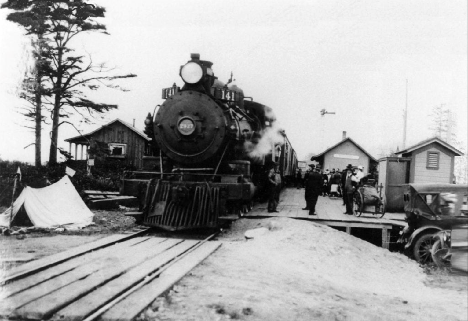 A steam locomotive parked at a small rural station, with nearby wooden buildings, a tent, and a parked car.