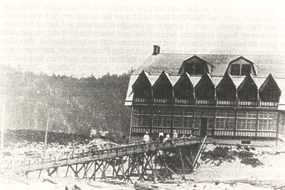 Black and white photo: Large wooden building with peaked roof, connected by a bridge. People are gathered on the bridge.