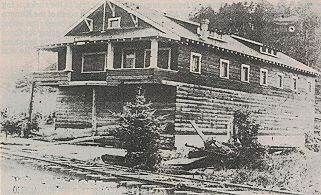 Lifted one story building sitting on log stilts laid parallel to the ground. Located near tracks and a few trees.