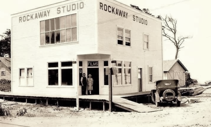 Rockaway Studio, two-story white building. Two people stand in doorway; old car parked nearby.