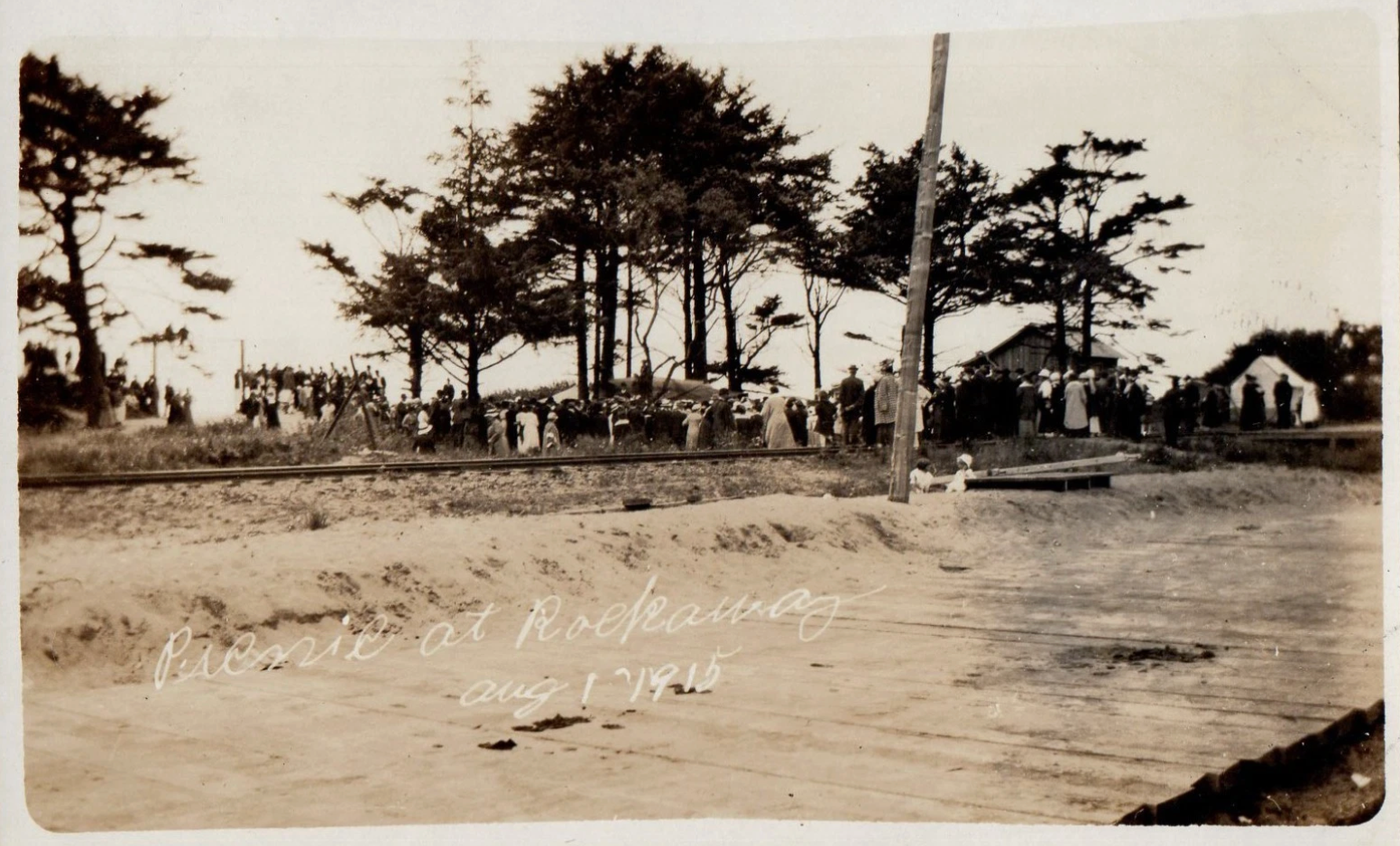 Sepia beach scene with people, tall trees, and small buildings along the shoreline