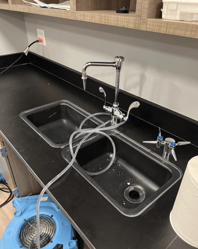 Black countertop with two sinks, faucet, and a connected hose near a floor fan drying after flood damage