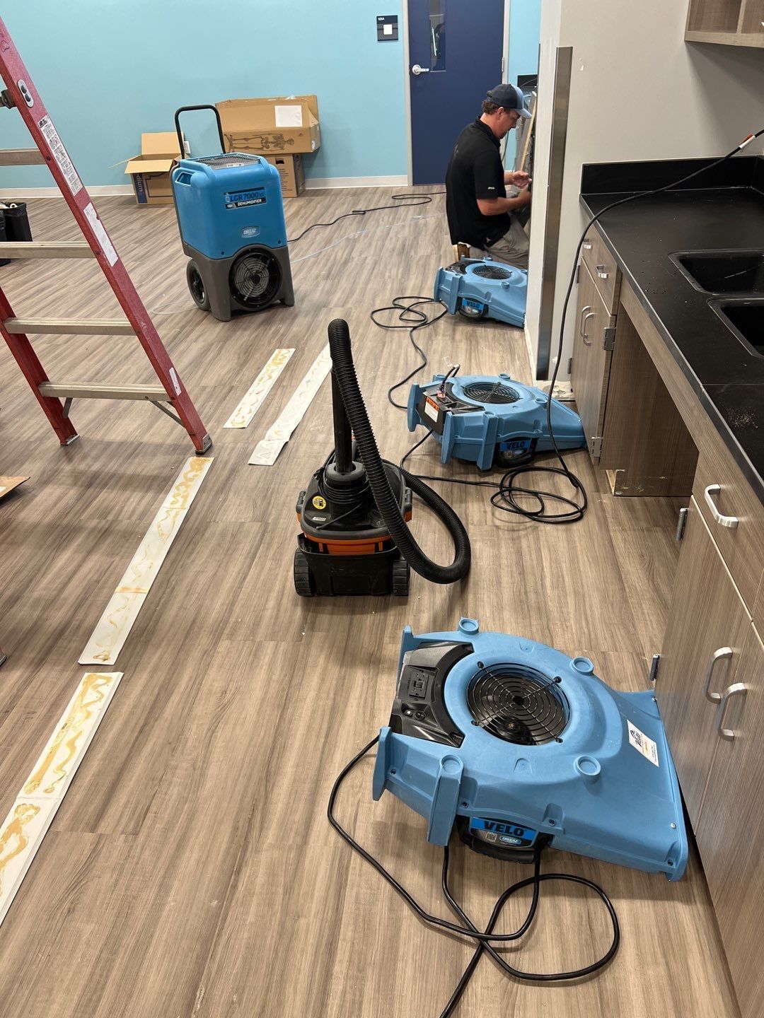 Workers drying a room with floor fans and a vacuum cleaner. One worker crouches near a dark cabinet.