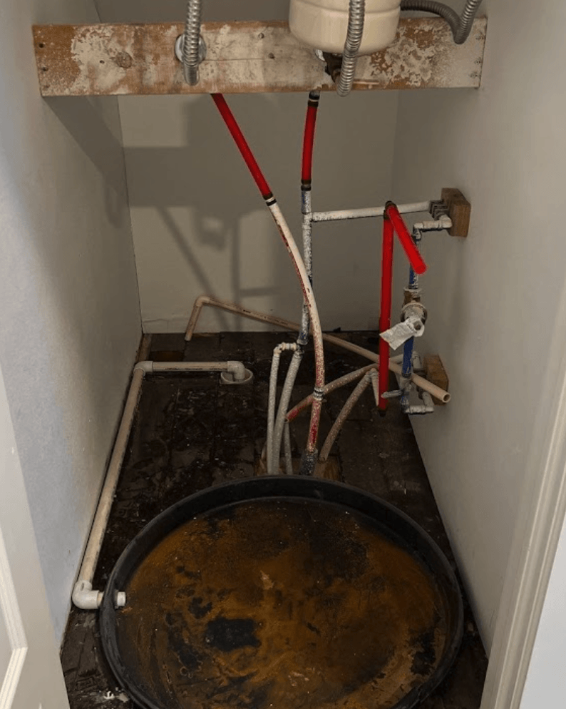 Plumbing in a dark closet with a black tub on the floor, red and white pipes, Water heater leak causing water damage