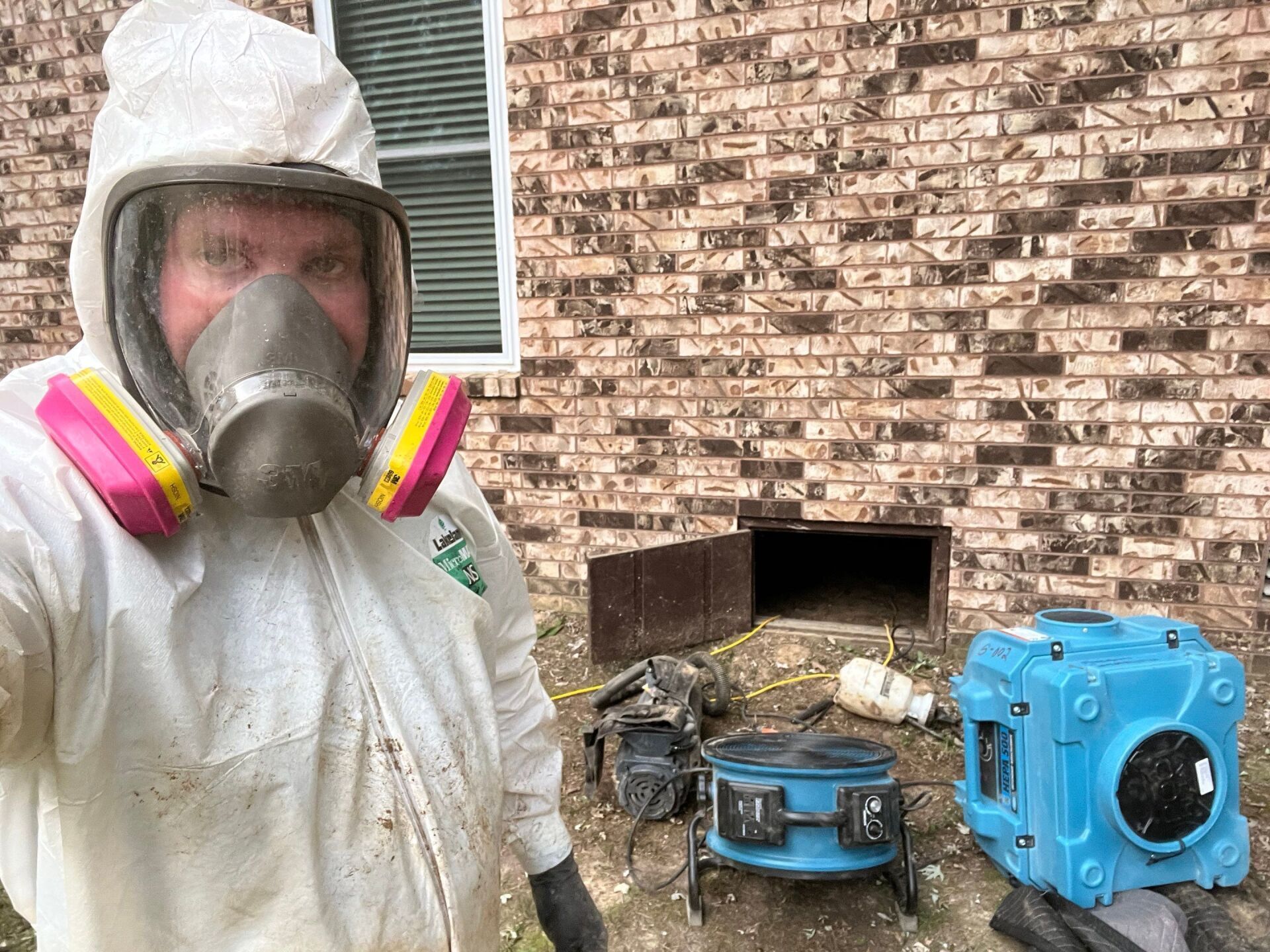 Person in protective suit and respirator by a brick wall and crawlspace entrance, with equipment.