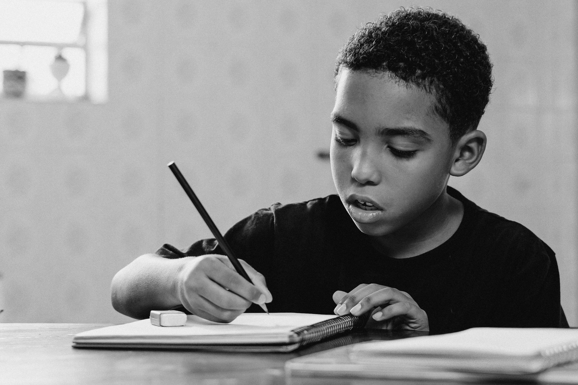 Boy writing in a notebook at a table, pencil in hand. Black and white photo.