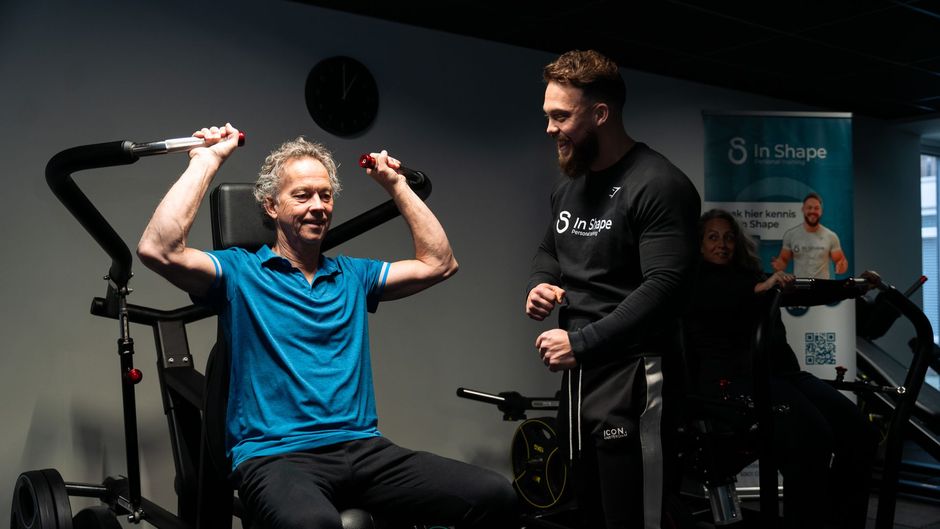 Een man tilt halters op in een sportschool terwijl een andere man toekijkt