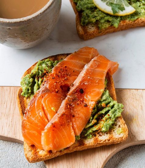 A close up of a sandwich with salmon and guacamole on a wooden cutting board.