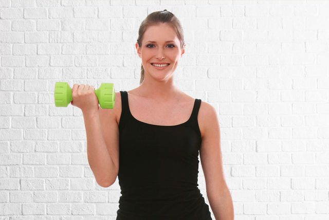 A woman is lifting a green dumbbell in front of a white brick wall.